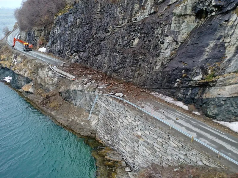 En vei mellom fjell og hav som er fotografert fra lufta. Deler av strekningen er ødelagt etter skred. Man kan se en gravemaskin lenger ned i veien. - Klikk for stort bilde