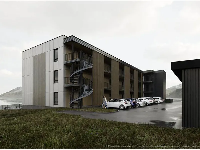 En moderne boligblokk med tre etasjer, sett fra veien. Spiraltrapp på byggets side, parkeringsplasser med biler foran. Utsikt mot sjøen og fjell i bakgrunnen.