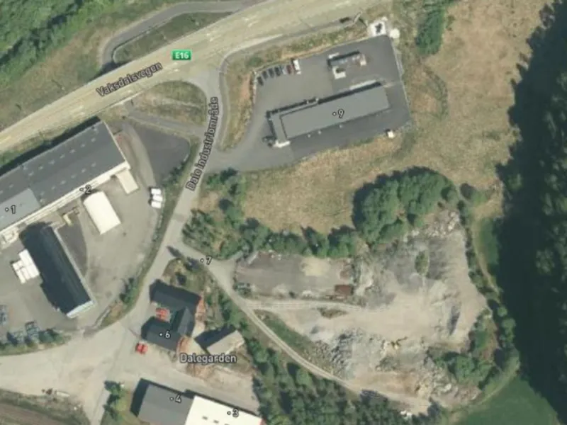 Satellittbilde som viser et industriområde med flere bygninger, veier og en åsside dekket av trær.