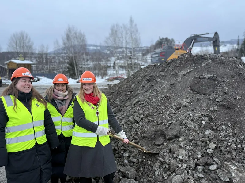 En gruppe mennesker med refleksvester og hjelmer foran en jordhaug og gravemaskin.