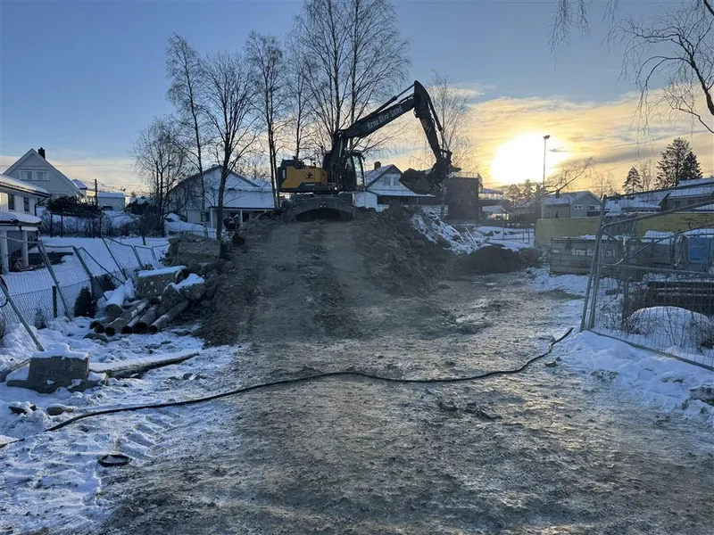 Gravemaskin på toppen av en jordfylling i et boligområde, med snø på bakken og sol som går ned bak husene. - Klikk for stort bilde