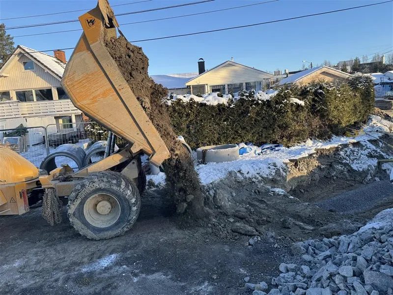 En anleggsmaskin tipper jord i en åpen grøft ved en boligvei, med snø, hekker og hus rundt anleggsområdet. - Klikk for stort bilde